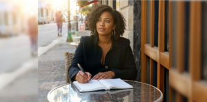 An elegant professional woman sitting at an outdoor cafe table, writing in an open journal to practice mindfulness and break free from autopilot living.